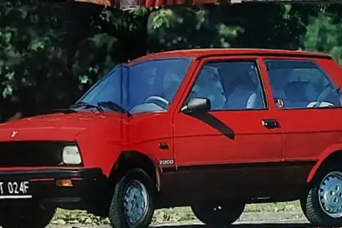 Czerwony yugo samochód z czarną atrapą chłodnicy i przyciemnianymi szybami, zaparkowany na tle zieleni.