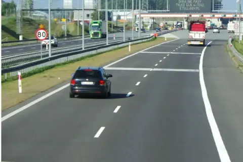 Czarny samochód jedzie autostradą, zbliżając się do bramek e-TOLL. Znak ograniczenia prędkości do 80 km/h przypomina o zachowaniu ostrożności.