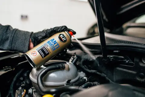 Ręka w rękawiczce rozpyla spray K2 Anti Marten pod maską samochodu, pokazując skuteczne rozwiązanie, czym odstraszyć kuny.