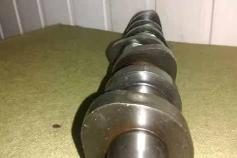 Metalowy wałek rozrządu z widocznymi śladami zużycia, noszący znamiona zatarty wałek rozrządu. Leży na zielonym materiale.