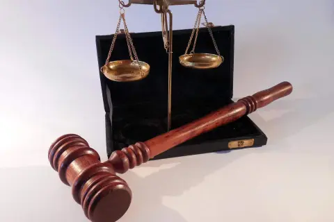 Młotek sędziowski i waga szalkowa leżą obok otwartego czarnego etui. Waga wygląda na lekko przechyloną, co może sugerować uszkodzenie mechaniczne lub nierównowagę.