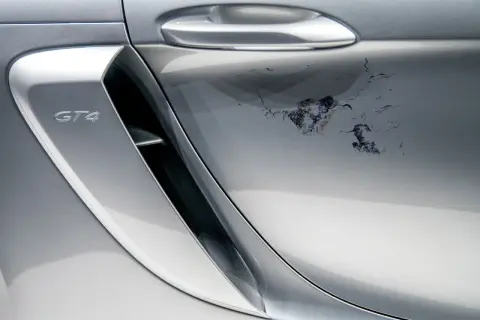Uszkodzony lakier bezbarwny na srebrnym Porsche GT4. Widoczne odpryski i zacieki wymagające naprawy.