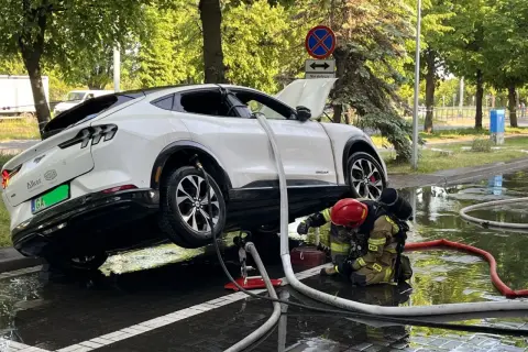Strażak w akcji, gaszenie samochodu elektrycznego. Woda i węże strażackie wokół białego SUV-a.
