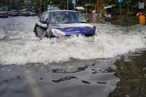 Niebieskie auto walczy z falą wody na zalanej ulicy. W tle widać inne auta po powodzi i autobusy.