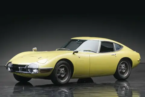 Żółta Toyota 2000 GT z lat 60. prezentuje swoje klasyczne, sportowe linie na błyszczącej podłodze.