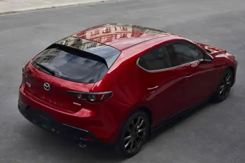 Czerwony Mazda 3 hatchback z czarnymi felgami, odbijający budynki. Elegancki, sportowy design, który odróżnia go od sedana.