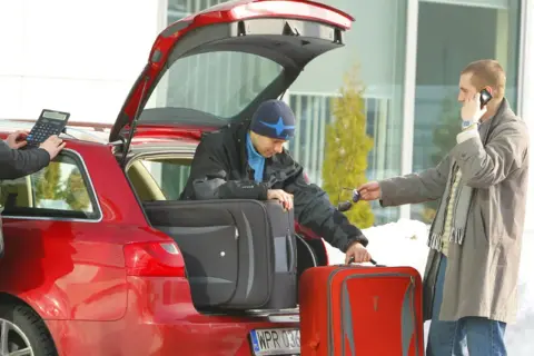 Rodzinne kombi gotowe do wyjazdu. Mężczyźni pakują bagaże, jeden sprawdza coś na kalkulatorze, drugi rozmawia przez telefon.
