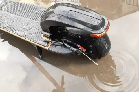 Czarna hulajnoga elektryczna stoi w kałuży. Czy hulajnoga elektryczna można jeździć w deszczu? Woda sięga do tylnego koła i części podestu.