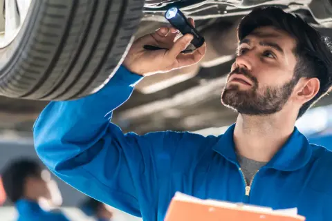 Mechanik w niebieskim kombinezonie oświetla latarką podwozie samochodu, wykonując **serwisowanie samochodu**.