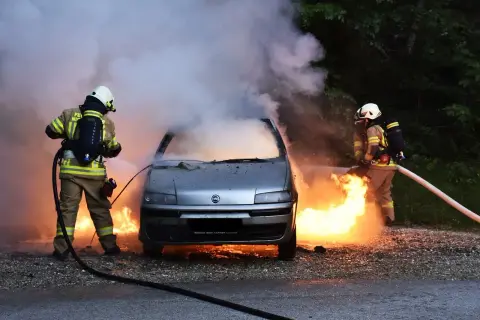 Strażacy walczą z płomieniami, gasząc samochód, który uległ samozapłonowi. Gęsty dym unosi się w powietrze.