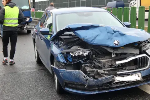 Widok na zniszczony przód niebieskiego samochodu po wypadku samochodowym. Na drodze widać osoby w kamizelkach odblaskowych.
