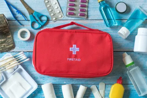 Czerwona torba z napisem FIRSTAID i krzyżem, obok niej wyposażenie apteczki samochodowej: plastry, bandaże, nożyczki, pęseta, leki, płyny.