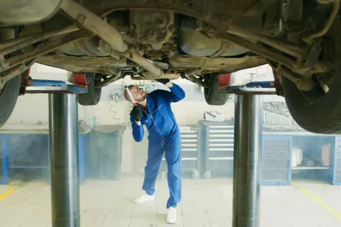 Mechanik w niebieskim kombinezonie bada podwozie samochodu na podnośniku. W powietrzu unosi się lekka mgiełka, przypominająca zapach benzyny w samochodzie.