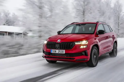 Czerwony crossover 4x4 pokonuje ośnieżoną drogę w zimowej scenerii, z domkiem w tle.