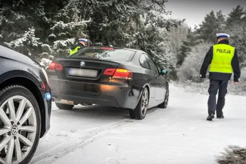 Policjant w kamizelce odblaskowej idzie w stronę czarnego BMW na zaśnieżonej drodze. Kierowca pewnie dostanie mandat za driftowanie.