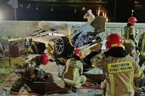Strażacy pracują przy wraku żółtego samochodu po wypadku Patryka Peretti. Auto jest mocno zniszczone, leży na boku.