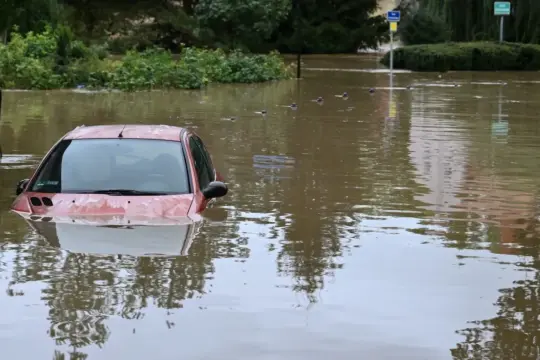 Czerwone auta po powodzi stoją zanurzone w brązowej wodzie. W tle widać drzewa i budynki.