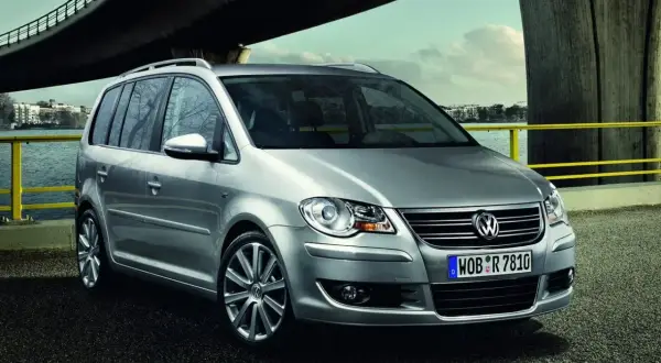Srebrny, bezawaryjny minivan Volkswagen Touran stoi nad wodą pod mostem.