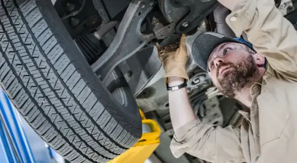 Mechanik w okularach ochronnych wykonuje sprawdzenie zawieszenia na stacji diagnostycznej. Widać jego rękę w rękawicy, która dotyka elementów podwozia.