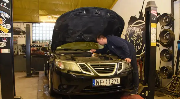 Mechanik sprawdza silnik czarnego Saaba. Na podnośniku warsztatowym, w tle widać narzędzia i części samochodowe.