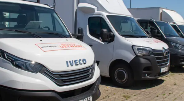 Rząd białych i ciemnych samochodów osobowo dostawczych zaparkowanych na zewnątrz. Na masce białego Iveco widnieje napis "Samochody Używane".