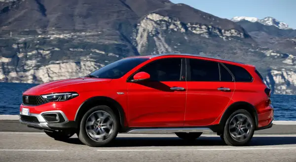 Czerwony Fiat Tipo Cross, wszechstronny samochód z segmentu kombi, przemierza malowniczą drogę nad jeziorem, z górami w tle.