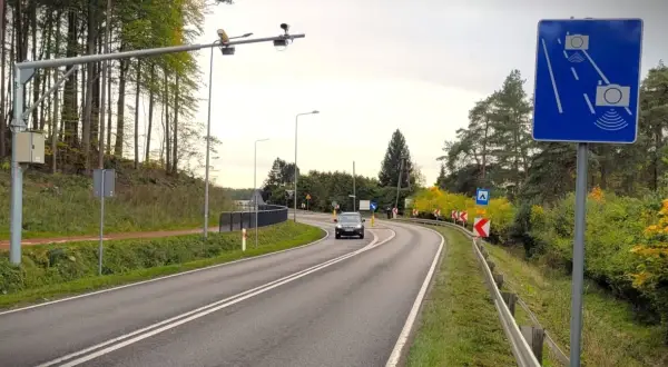 Znak informuje o systemie do pomiaru prędkości w nocy przez policję. Droga kręta, otoczona drzewami.