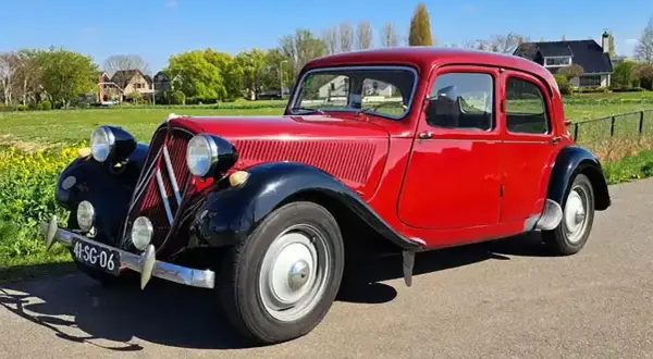 Klasyczny, czerwony Citroen Traction Avant, jeden z kultowych modele stare, zaparkowany na tle zielonej łąki i domów.