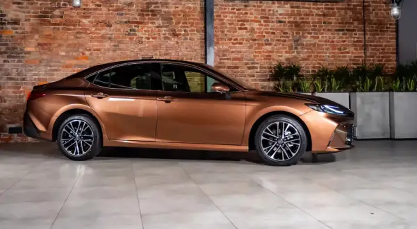 Brązowy sedan o eleganckiej linii, jeden z najładniejszych sedanów, prezentuje się na tle ceglanej ściany i zieleni.