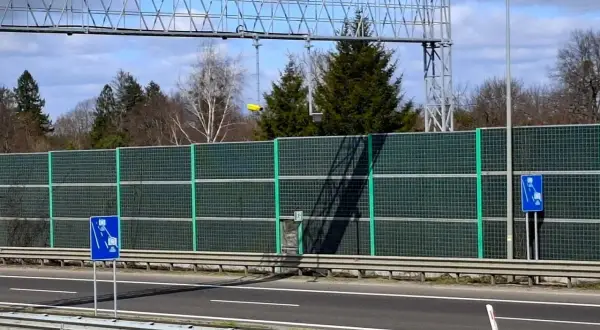 Znak informuje o odcinkowym pomiarze prędkości. Za zieloną barierą dźwiękochłonną widać drzewa i niebo.