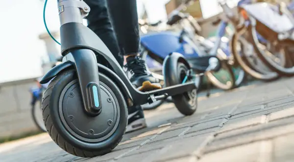 Osoba na hulajnodze elektrycznej, gotowa do jazdy. W tle stoją rowery miejskie. Przepisy hulajnoga elektryczna to ważna sprawa.