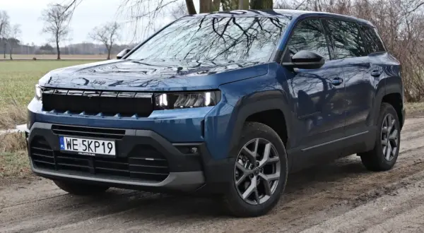 Niebieski SUV klasy premium Jeep Compass na błotnistej drodze, z drzewami w tle.