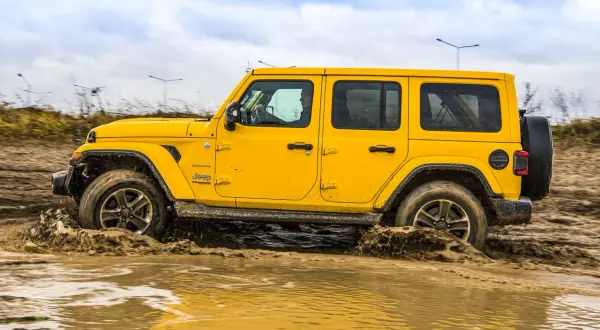 Żółty Jeep Wrangler Sahara pokonuje błotniste rozlewiska, pokazując swoje możliwości terenowe 4x4.
