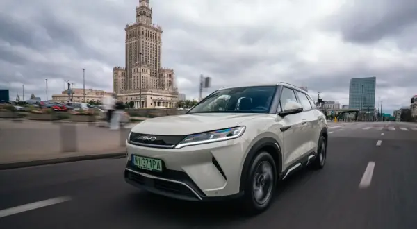 Biały SUV z logo chińskich samochodów BYD jedzie ulicą w Warszawie, z Pałacem Kultury w tle.