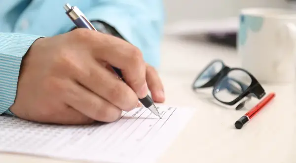 Dłoń pisze na dokumencie, obok okulary i kubek. Zastanawiasz się, co to znaczy RRSO? To roczna rzeczywista stopa oprocentowania.
