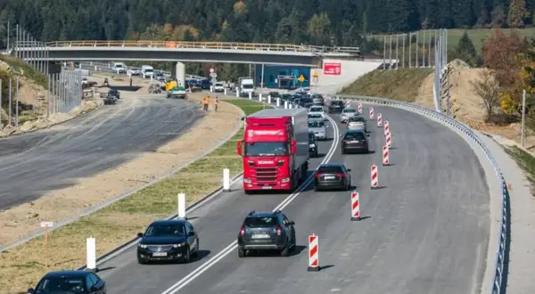 Budowa zakopianki w otoczeniu lasu. Ruch samochodowy, w tym czerwona ciężarówka, odbywa się po nowej drodze z mostem w tle.
