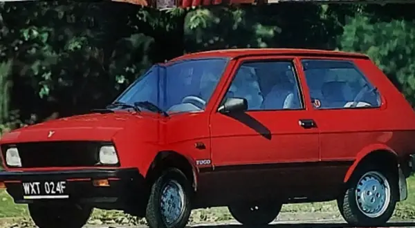 Czerwony yugo samochód z czarną atrapą chłodnicy i przyciemnianymi szybami, zaparkowany na tle zieleni.