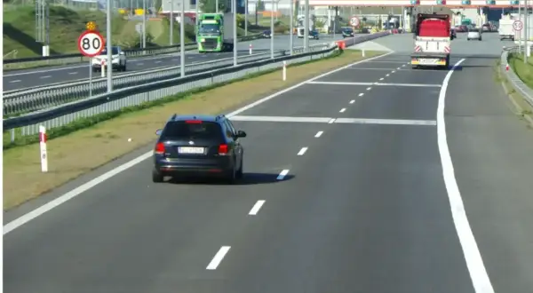 Czarny samochód jedzie autostradą, zbliżając się do bramek e-TOLL. Znak ograniczenia prędkości do 80 km/h przypomina o zachowaniu ostrożności.