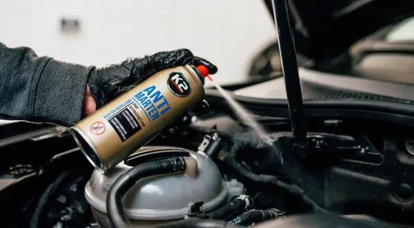 Ręka w rękawiczce rozpyla spray K2 Anti Marten pod maską samochodu, pokazując skuteczne rozwiązanie, czym odstraszyć kuny.
