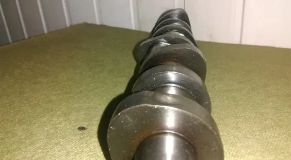 Metalowy wałek rozrządu z widocznymi śladami zużycia, noszący znamiona zatarty wałek rozrządu. Leży na zielonym materiale.