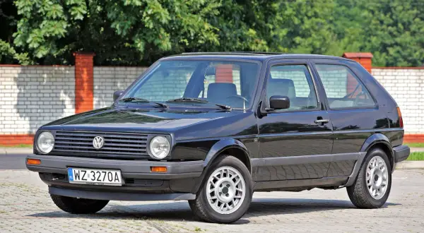 Czarny VW Golf 2, klasyk z lat 80., lśni w słońcu na tle ceglanego muru i zieleni.