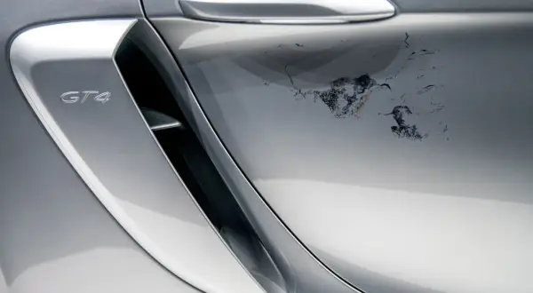 Uszkodzony lakier bezbarwny na srebrnym Porsche GT4. Widoczne odpryski i zacieki wymagające naprawy.