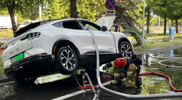 Strażak w akcji, gaszenie samochodu elektrycznego. Woda i węże strażackie wokół białego SUV-a.