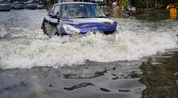 Niebieskie auto walczy z falą wody na zalanej ulicy. W tle widać inne auta po powodzi i autobusy.