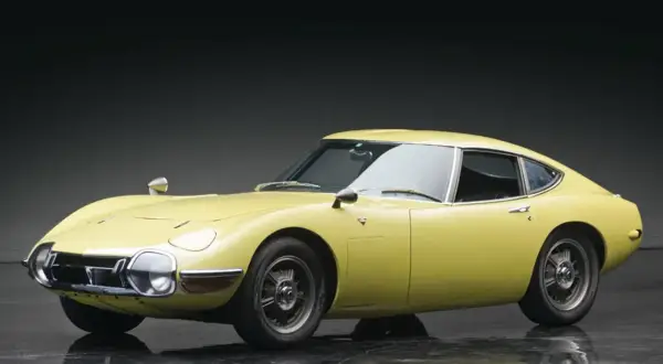Żółta Toyota 2000 GT z lat 60. prezentuje swoje klasyczne, sportowe linie na błyszczącej podłodze.