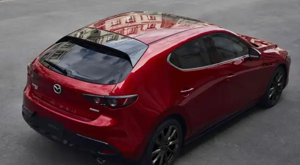 Czerwony Mazda 3 hatchback z czarnymi felgami, odbijający budynki. Elegancki, sportowy design, który odróżnia go od sedana.