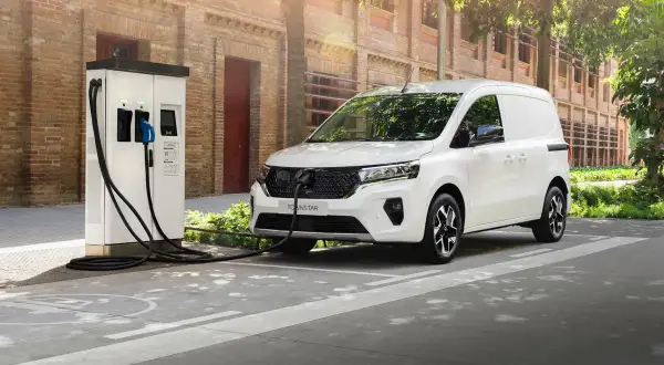 Biały samochód dostawczy podłączony do ładowarki elektrycznej na tle ceglanego budynku.