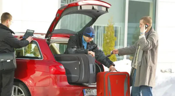 Rodzinne kombi gotowe do wyjazdu. Mężczyźni pakują bagaże, jeden sprawdza coś na kalkulatorze, drugi rozmawia przez telefon.