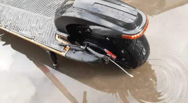 Czarna hulajnoga elektryczna stoi w kałuży. Czy hulajnoga elektryczna można jeździć w deszczu? Woda sięga do tylnego koła i części podestu.
