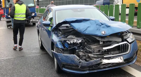 Widok na zniszczony przód niebieskiego samochodu po wypadku samochodowym. Na drodze widać osoby w kamizelkach odblaskowych.